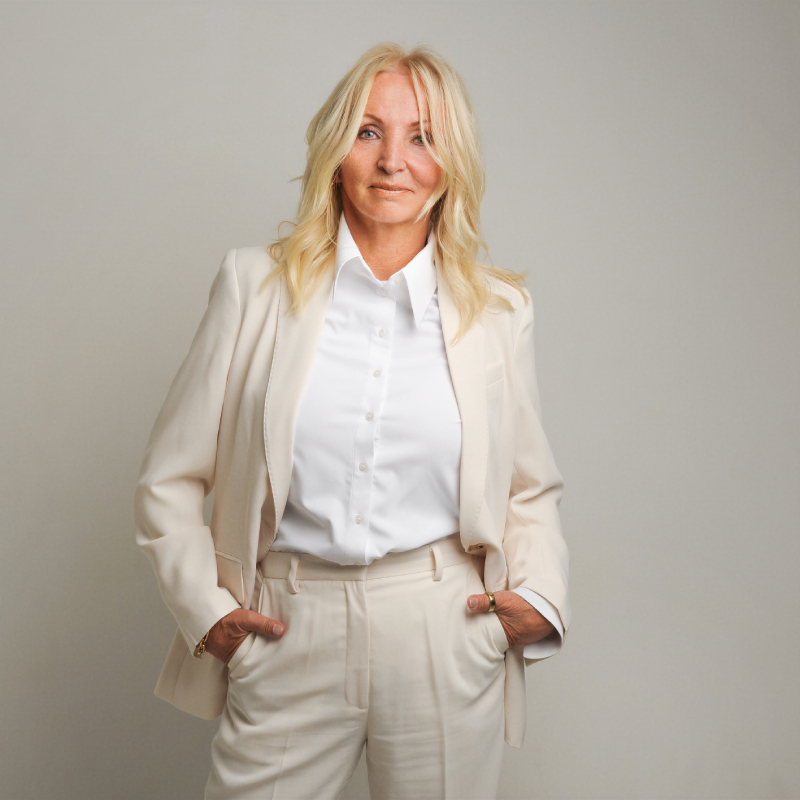 A woman with long blonde hair wearing a light-colored suit and a white shirt stands confidently with her hands in her pockets.